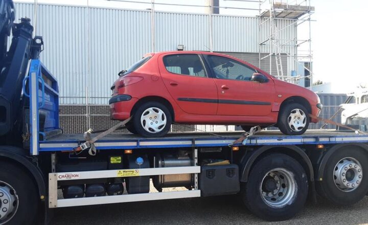 A red car strapped onto a flatbed truck near an industrial building.