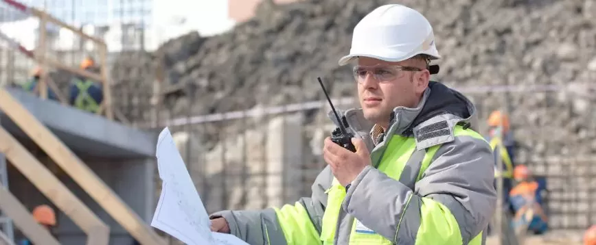 A construction worker in safety gear holds a blueprint and uses a walkie-talkie.