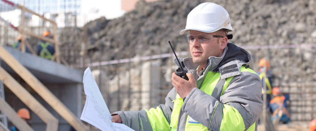 Construction worker in reflective jacket and helmet holding plans and a walkie-talkie outside.