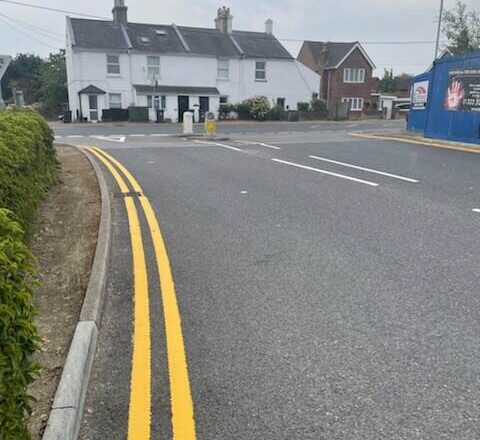Road with double yellow lines, hedges, white buildings, and a blue hoarding on the corner.