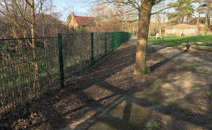 A green wire fence alongside a quiet pathway with trees and buildings nearby.