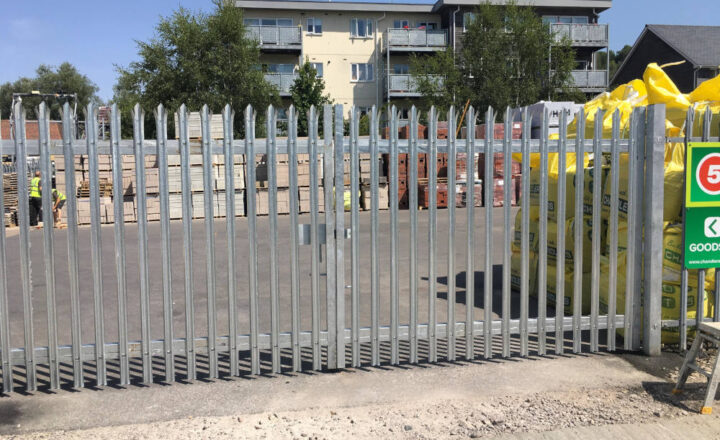 Metal fence with security spikes surrounds a construction site with supplies and a warning sign.