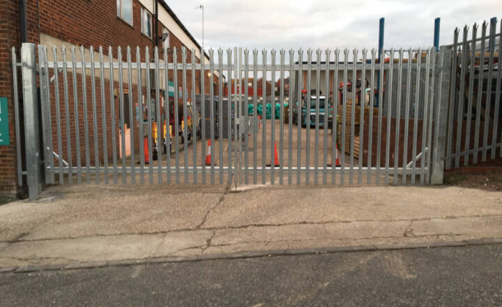 Metal security gate surrounds an industrial area with traffic cones and parked vehicles visible.