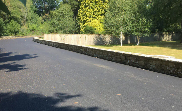 A newly paved road curves alongside a stone wall, flanked by trees and grass.