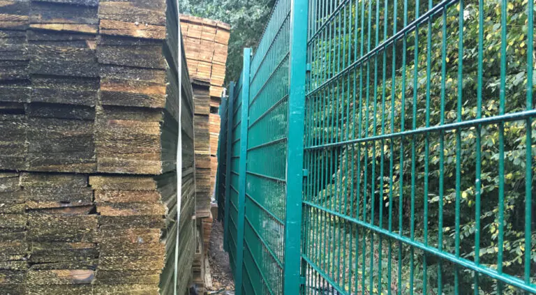 Stacks of wooden boards on the left, green metal fence on the right.