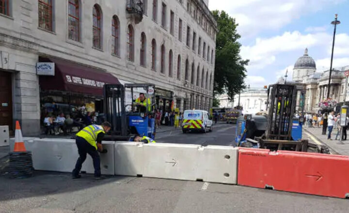 Workers install barriers on a street near a historic building and an emergency vehicle.