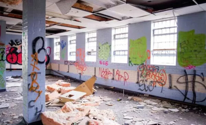 Abandoned room with graffiti-covered walls, broken ceiling tiles, and debris scattered on the floor.