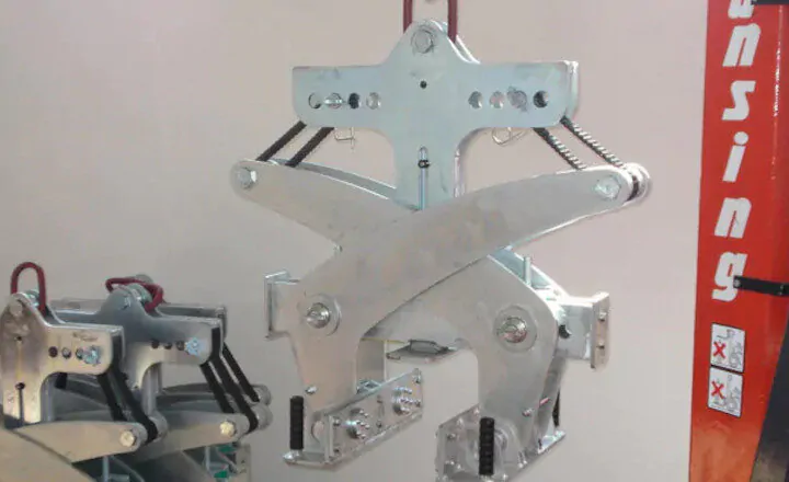 Suspended industrial metal clamp mechanism with springs, mounted on an orange vertical structure.
