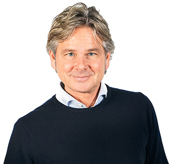 A man smiles, wearing a black jumper and white shirt.