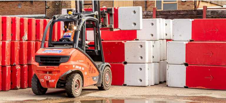 A forklift transports a stack of red and white concrete blocks outdoors.