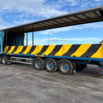 Large flatbed truck transporting yellow and black striped traffic barriers with the trailer curtain open.