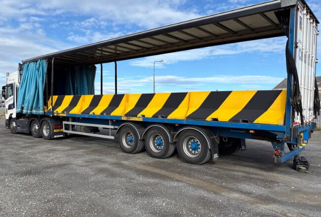 Large flatbed truck transporting yellow and black striped traffic barriers with the trailer curtain open.